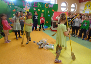Dzieci śpiewają piosenkę, na środku tańczą trzy dziewczynki z miotłami wokół rozsypanych odpadków.